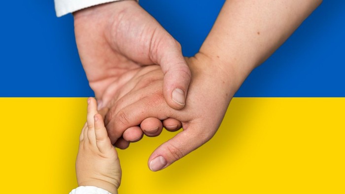 En voksens og et barns hånd holder hinanden foran en blå og gul flag.