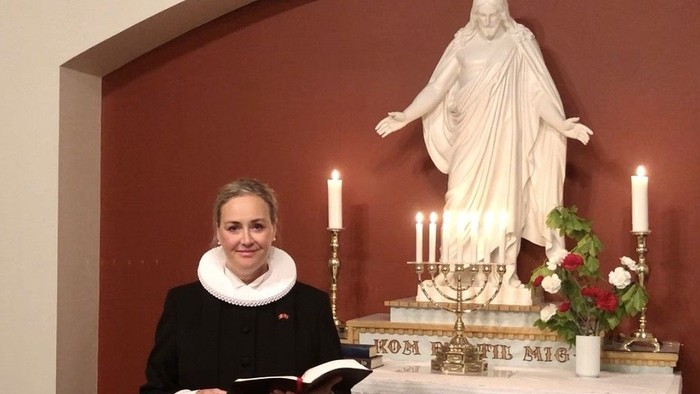 Kvinne i mørkt tøj holder en bog og står foran alter med Jesus-statue og lysede lygter