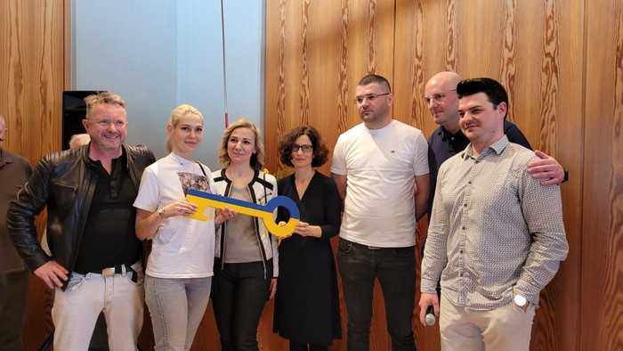 Eine Gruppe von sieben Personen steht vor einer Holzwand, eine hält eine Gitarre