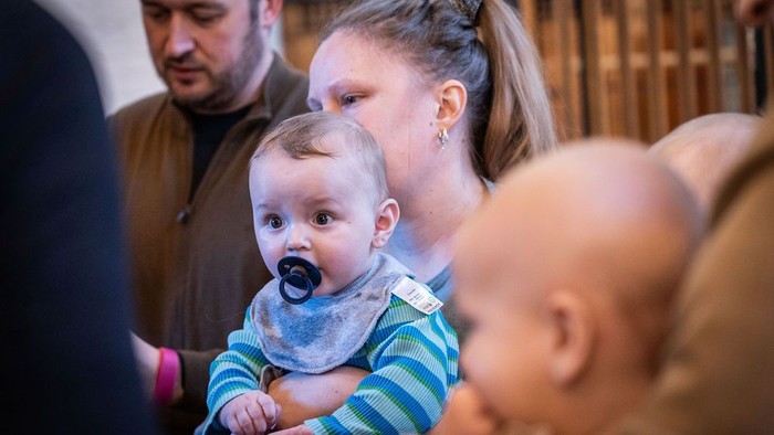 En kvinde holder en baby med en sut, mens en anden baby er i nærheden, og en mand står bag dem.