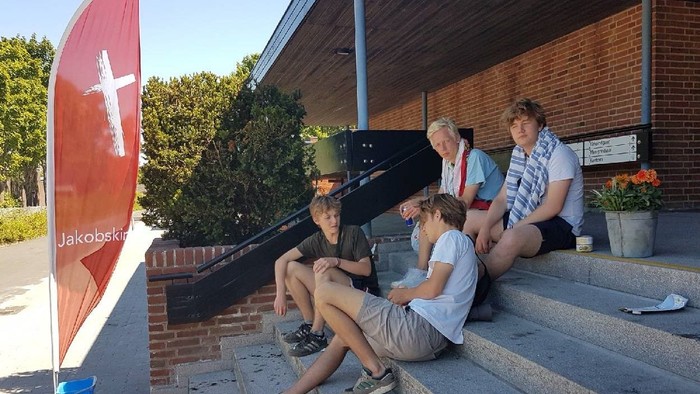 En gruppe mennesker sidder på træer foran et rødt bjærgede hus med en flagstang med et kors.