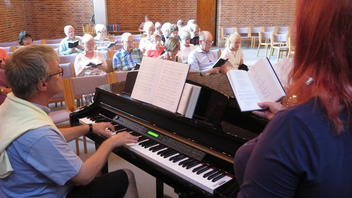 En gruppe af mennesker deltager i en sangprove, hvor en mand spiller piano og en kvinde holder partituret op.
