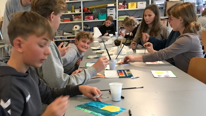 Børn og unge sidder ved bord og maler med vandsmalerier i en klassesal.