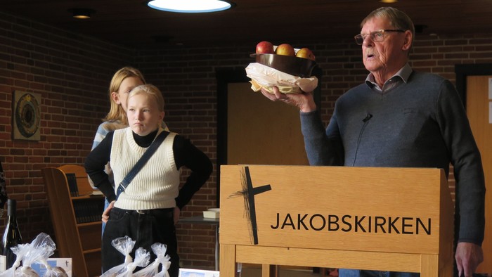 En mand holder et brød op for at vise, mens en kvinde og et barn står ved siden af. Der er et bord med brød bag dem.