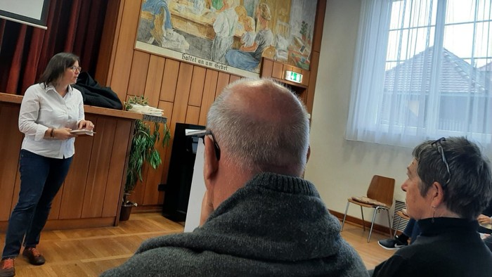 Eine Frau steht vor einem großen Wandteppich und hält eine Rede, während zwei Männer zuhören.