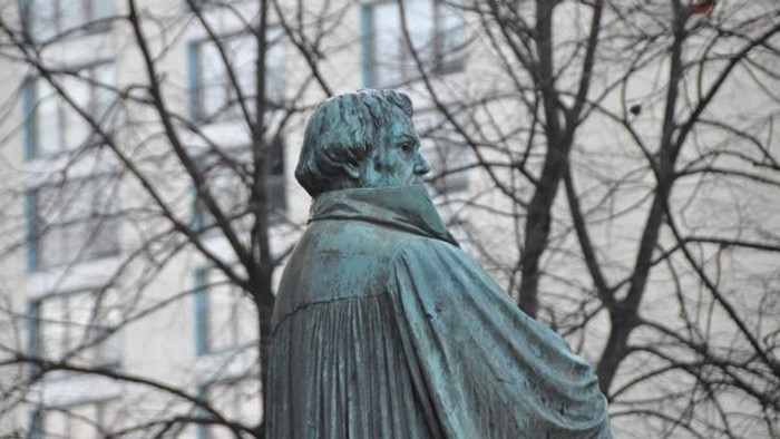 Die Bronzestatue eines Mannes mit Umhang vor Bäumen und Gebäude.