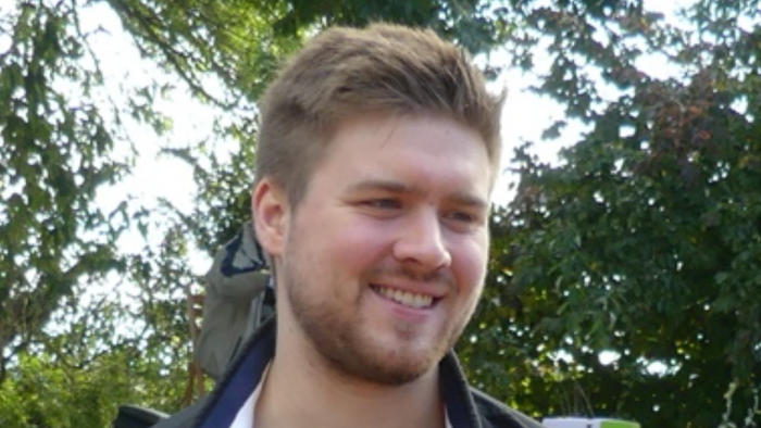 Ein junger Mann lächelt im Freien vor Bäumen.