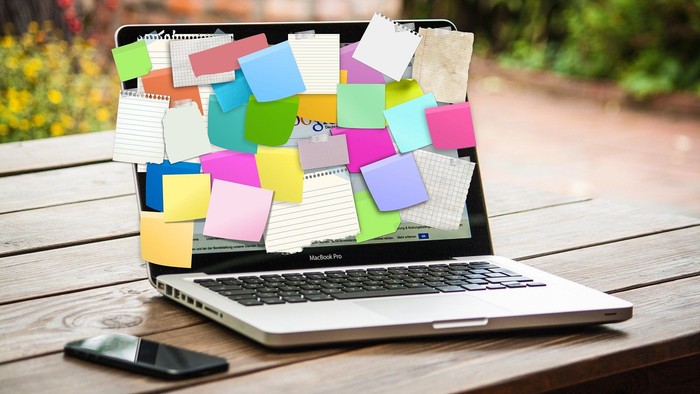 En MacBook Pro dekket af mange farvede post-it noter, placeret på en træbæn ved siden af et mobiltelefon.
