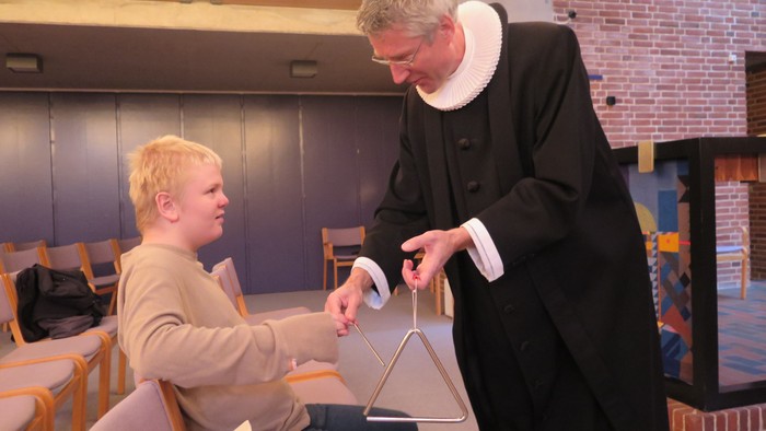 En præst hjælper en ung dreng med at spille på en cymbaler i en kirke.