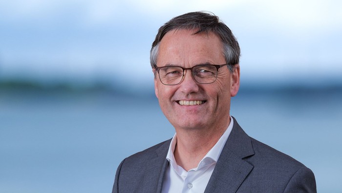 Ein Mann in Anzug und Brille lächelt vor einem Wasserhintergrund.