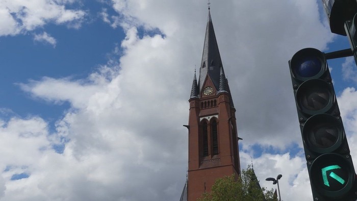 Ein roter Kirchturm mit spitzem Dach vor blauem Himmel und Wolken, daneben eine Ampel.