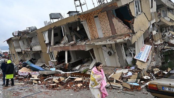 Ein zerstörtes Gebäude nach einem Erdbeben, Menschen in Sicherheitswesten und eine Frau in Decke stehen vor den Trümmern.