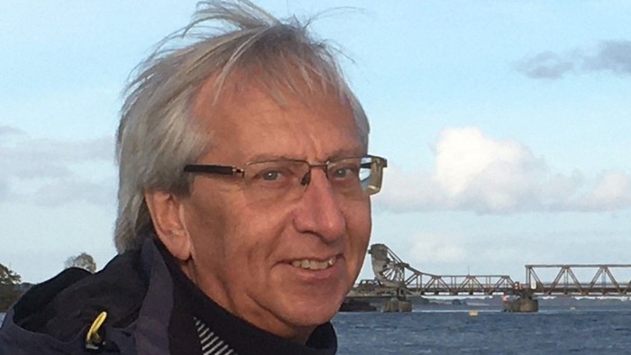 Ein älterer Mann mit Brille lächelt vor einem Wasserlauf und einer Brücke.