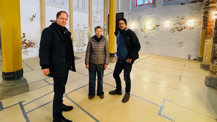 Drei Männer stehen in einem Raum mit Säulen und hohen Fenstern.