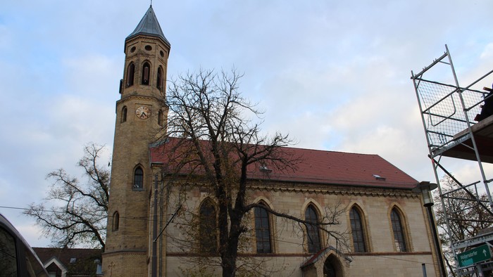 Altes Kirchengebäude mit hohem Turm und spitzen Dach, umgeben von kahlen Bäumen und einem grauen Himmel.