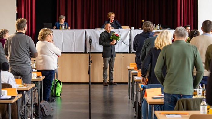Eine Gruppe von Menschen hört einem Mann mit Blumenstrauß auf einer Bühne zu.