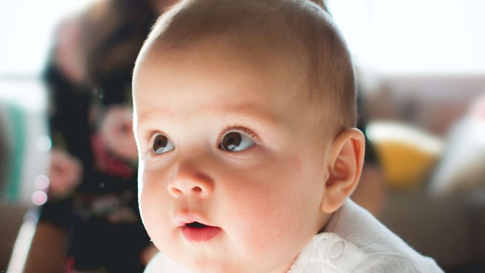 Et spæd barn i hvidt tøj ser opad, mens en kvinde smiler i baggrunden