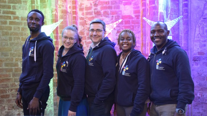 Eine Gruppe von fünf Personen in blauen Kapuzenpullovern steht vor einer farbenfrohen Ziegelwand.