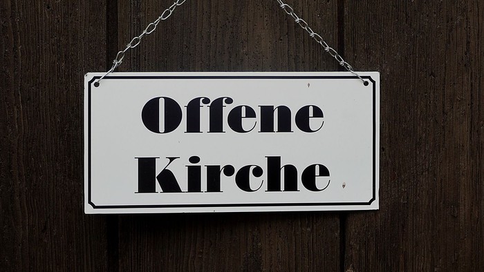 Ein Schild mit der Aufschrift *„Offene Kirche“* hängt an einer Holzwand.