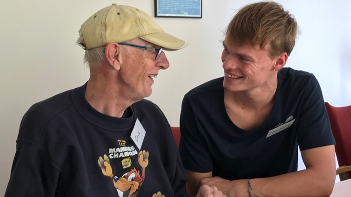 To oldere mænd sidder ved bord og smiler, en bog og en tasse på bordet
