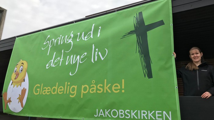 En kvinde står ved et grønt banner med påskesalutationer og et kors