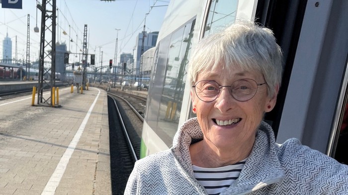 Eine ältere Frau steht lachend an einem Bahnhof neben einem Zug.