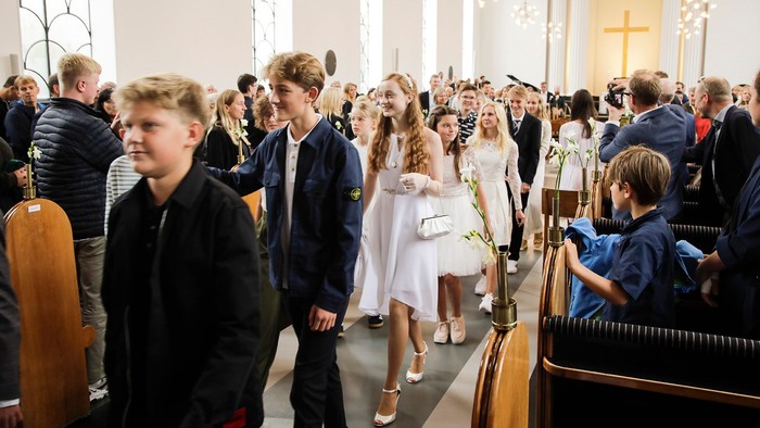 En kirkefuld med børn og voksne, nogle står, nogle sidder, alle er formelt klædt.