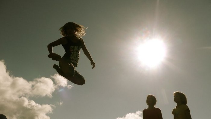 Ein Kind springt hoch in die Luft, während zwei andere Kinder zusehen und die Sonne scheint hell.