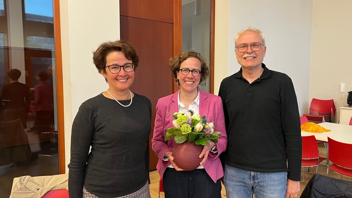 Drei Personen stehen in einem Raum, eine Frau hält Blumen