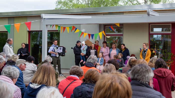 Eine Gruppe von Menschen versammelt sich vor einem Gebäude mit bunten Fahnen, einige stehen auf einer Bühne.