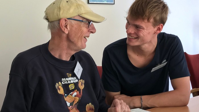 To oldere mænd sidder ved bord og smiler, en bog og en tasse på bordet