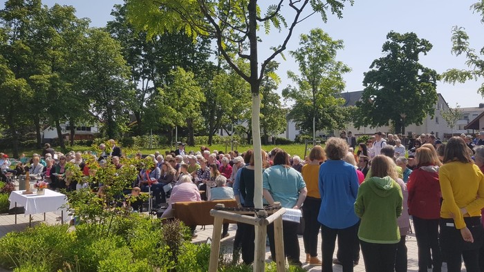 Eine Gruppe von Menschen versammelt sich im Freien, einige stehen, andere sitzen an Tischen, umgeben von Bäumen und Gebäuden.