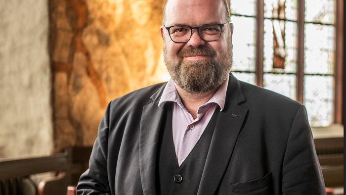 Mann mit Brille und Bart posiert in Sakko vor steinerner Wand.