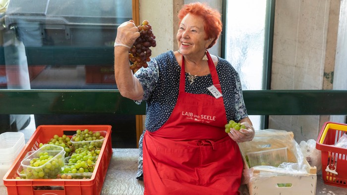 Eine fröhliche Frau in einer roten Schürze hält Trauben, während sie an einem Marktstand mit Obst und Gemüse arbeitet.