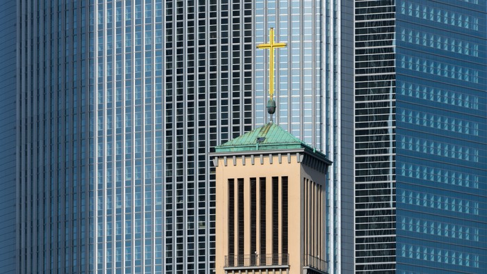 Ein gelber Kirchturm mit grünem Dach und Kreuz zwischen zwei modernen Wolkenkratzern.