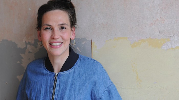 Eine Frau in blauer Jacke lächelt vor einer abblätternden Wand.