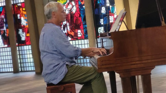 Ein älterer Mann spielt Klavier in einem Raum mit farbenfrohen Gemälden.