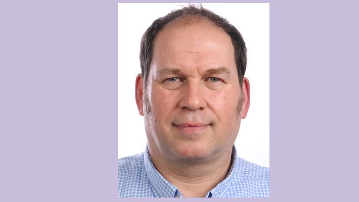 Ein Mann mit dunklem Haar und einem blauen Hemd lächelt in die Kamera.