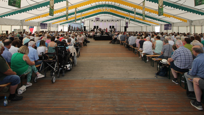 Große Menschenmenge sitzt in einem Zelt und hört einem Redner zu.