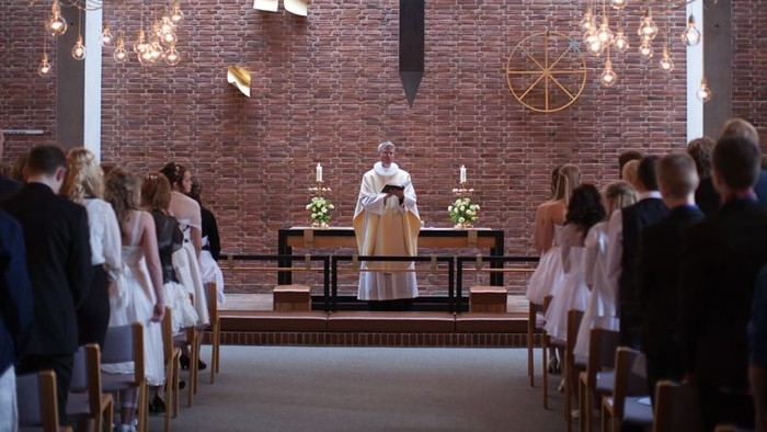 En præst udfører en ceremoniel handling i en moderne kirke med røde mursten og hængende lygter.