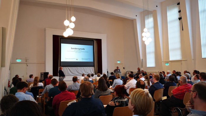 Vortrag in einem großen Saal mit vielen Zuhörern und Projektionsbildschirm