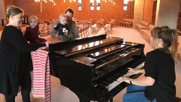 En gruppe børn og en voksne kvinde spiller på et sort piano i et rum med mursten og hængelys.