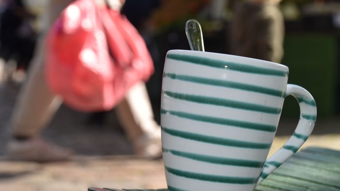 Eine Tasse mit grün-weißen Streifen steht auf einem Holztisch, im Hintergrund sitzt jemand in einem roten Kleid.