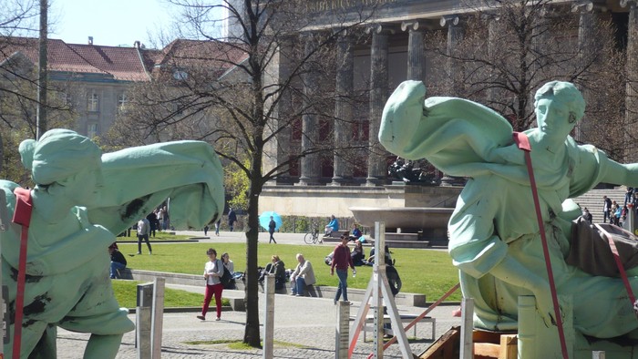 Große grüne Statuen auf einem Platz vor einem Gebäude mit Säulen, Menschen im Hintergrund