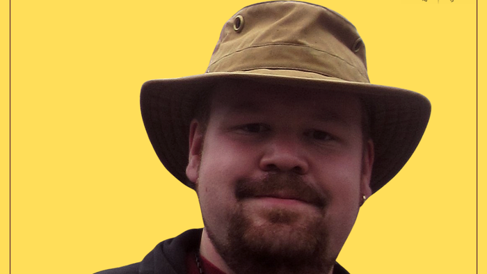 Ein Mann mit Hut und Kreuzanhänger vor gelbem Hintergrund