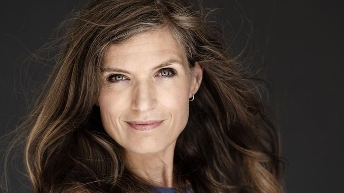 En kvinde med langt, brunt hår, skærer armene og smiler til kameraet.