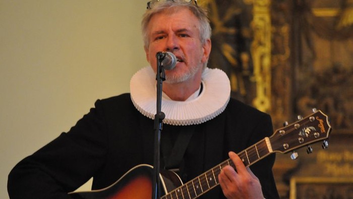 En mand spiller guitar og synger i et mørkt tøj med hvid krage.