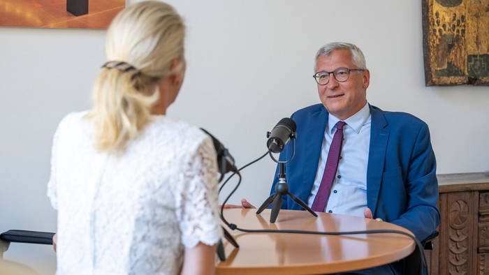 Ein Mann in Anzug sitzt an einem Tisch mit Mikrofon, eine Frau steht neben ihm