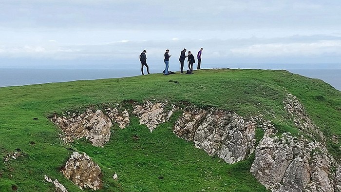 Fünf Personen stehen auf einem grasbewachsenen Hügel mit Felsen und Blick auf das Meer.