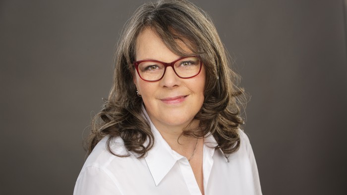 Eine Frau mit Brille und welligem Haar, die ein weißes Hemd trägt.